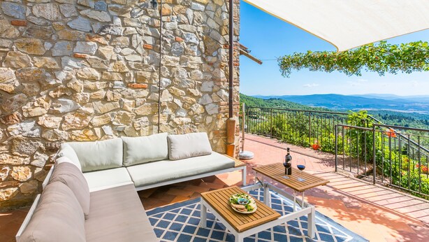 A stone wall, a white sofa, a wooden table with a plate, and a view of a vineyard.