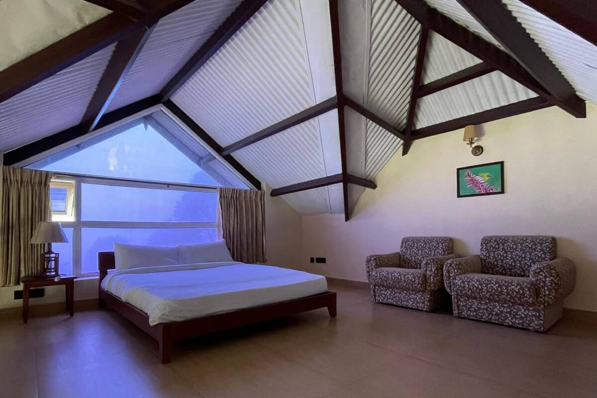 Een slaapkamer met een groot raam, een bed, twee gestreepte fauteuils en een kleine tafel met een lamp.