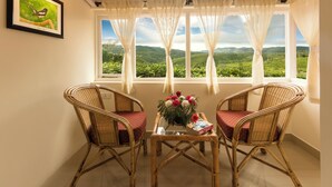 A room with two wicker chairs, a small table with a vase of flowers, and a framed picture on the wall. - Teanest Nightingale - Nature Resorts (Kotagiri)