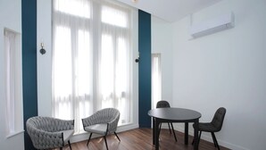 A room with a round table and four chairs, large windows with sheer curtains, and a white air conditioning unit on the wall.