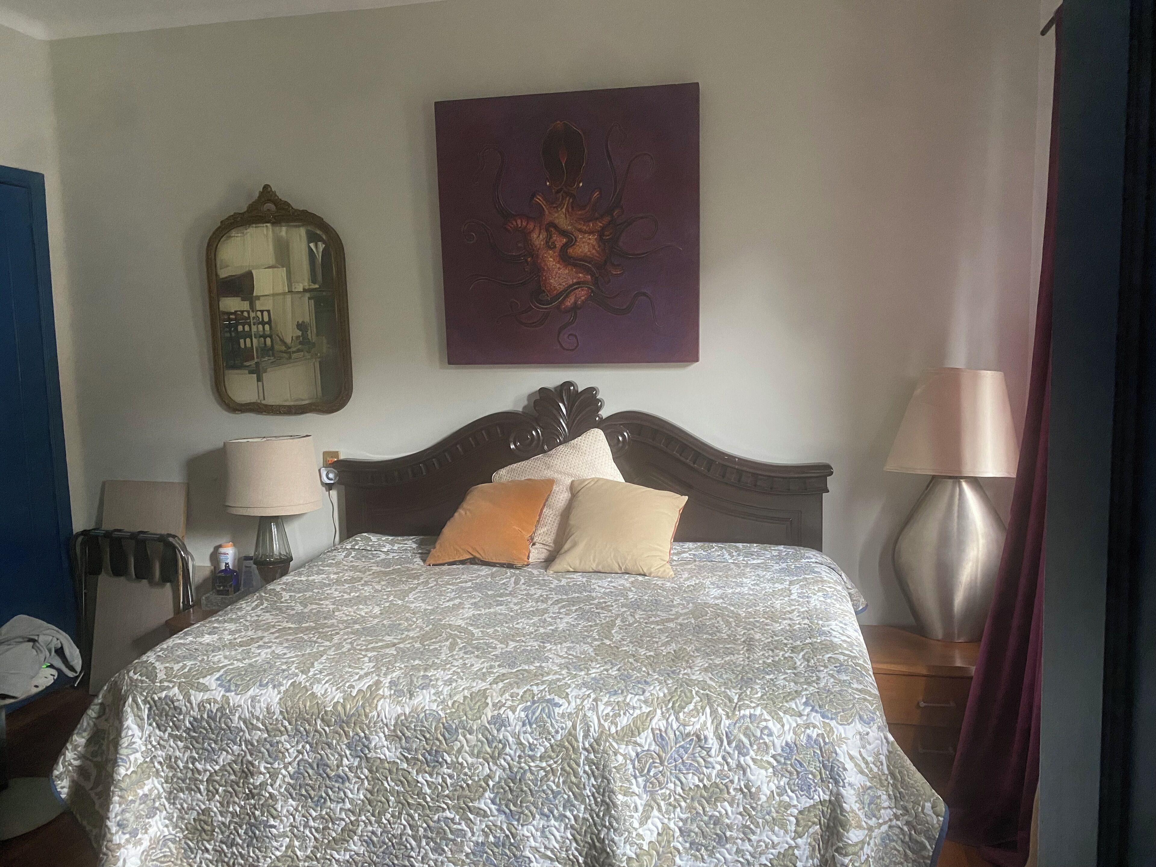 A neatly made bed with a patterned bedspread, a mirror, a painting, and a lamp on a bedside table.