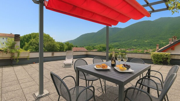 A terrace with a table set for two, a red umbrella, and a scenic mountain view.
