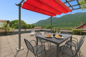 A terrace with a table set for two, a red umbrella, and a scenic mountain view.