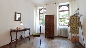 A room with a wooden dresser, a chair, a table with a lamp, a mirror, and a clothing rack.