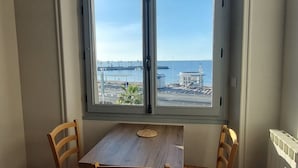 Un coin repas avec une table et quatre chaises, une grande fenêtre en arc de cercle offrant une vue sur la mer et les bateaux, et un luminaire suspendu.