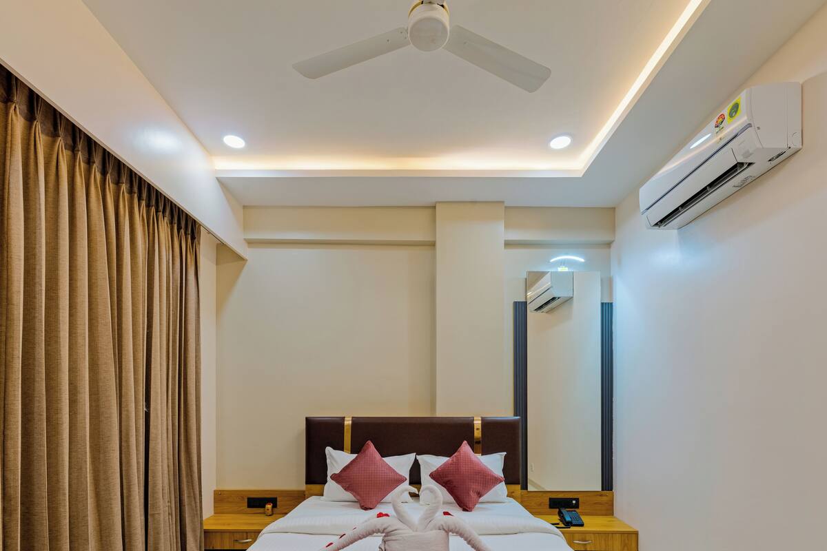 A hotel room with a bed, curtains, a ceiling fan, an air conditioning unit, and a bedside table with a phone.