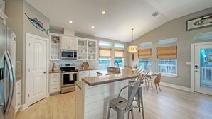 A modern kitchen with white cabinetry, a central island, bar stools, and a dining area with a view of the outside. - Sleeps 24 | 2 Homes w/ Beach Boardwalk Access & Deck Views | Steps to the Beach (Port Aransas)