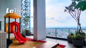 Un espace de jeux pour enfants avec un toboggan rouge, un banc et des plantes en pot, situé sur un balcon offrant une vue sur la ville.