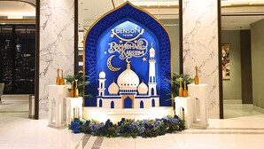 Une arche bleue sur laquelle on peut lire « Benson Tower » et « Ramadhan Kareem », entourée de plantes et de fleurs décoratives.