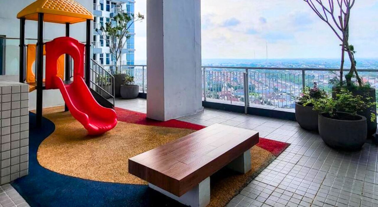 Area giochi per bambini con uno scivolo rosso, una panchina e piante in vaso su un balcone con vista sulla città.