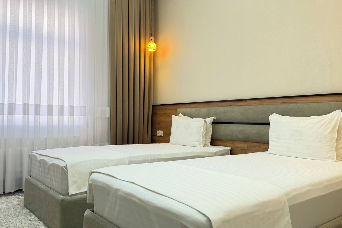 Een hotelkamer met twee bedden, een houten hoofdeinde, een wandlamp en dunne gordijnen.