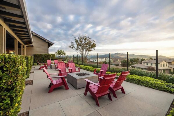 Un patio al aire libre con sillas Adirondack rojas y un brasero, rodeado de follaje y con vistas a una zona residencial.