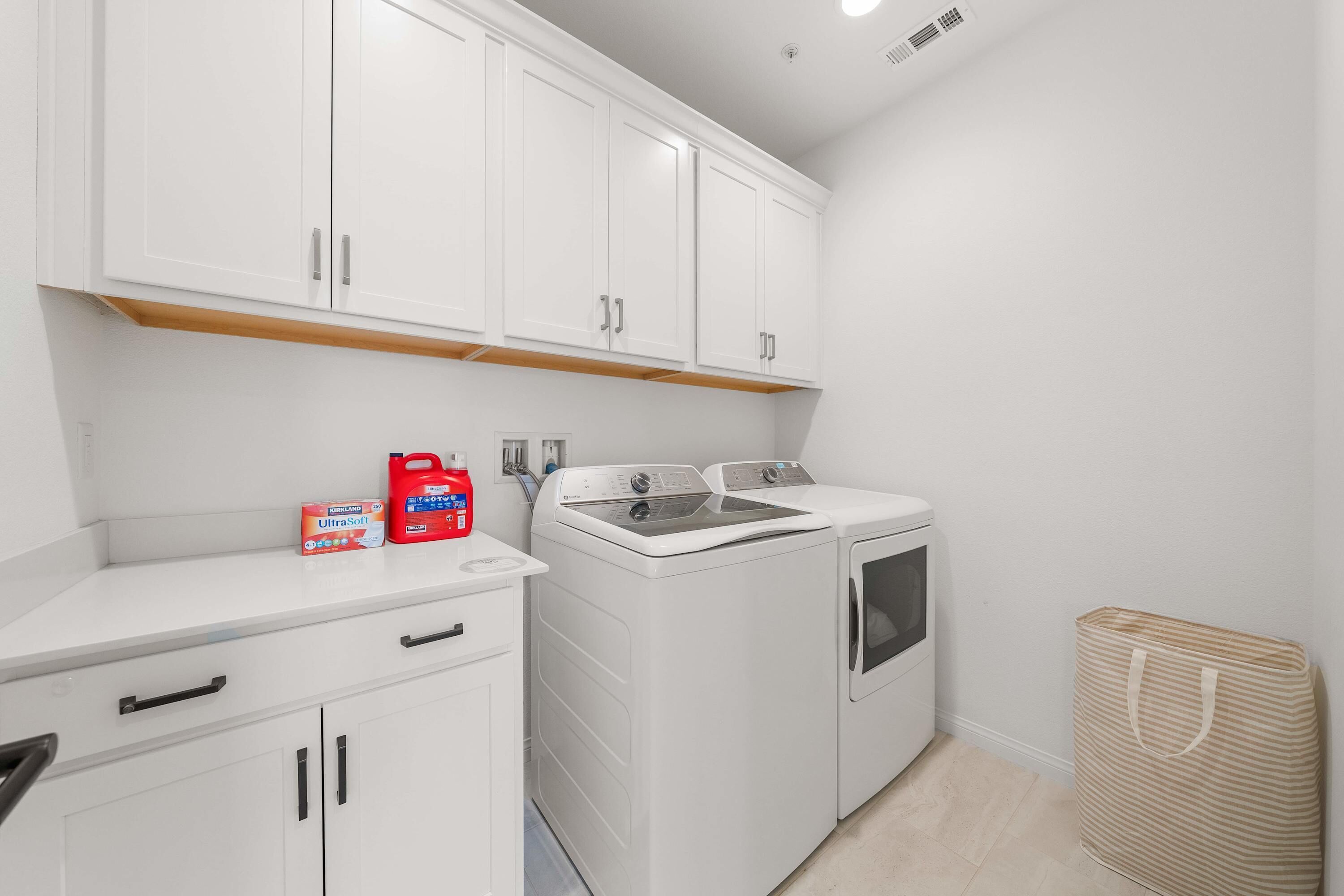 A laundry room with white cabinets, a washing machine, and a dryer. There are detergent bottles on the counter.