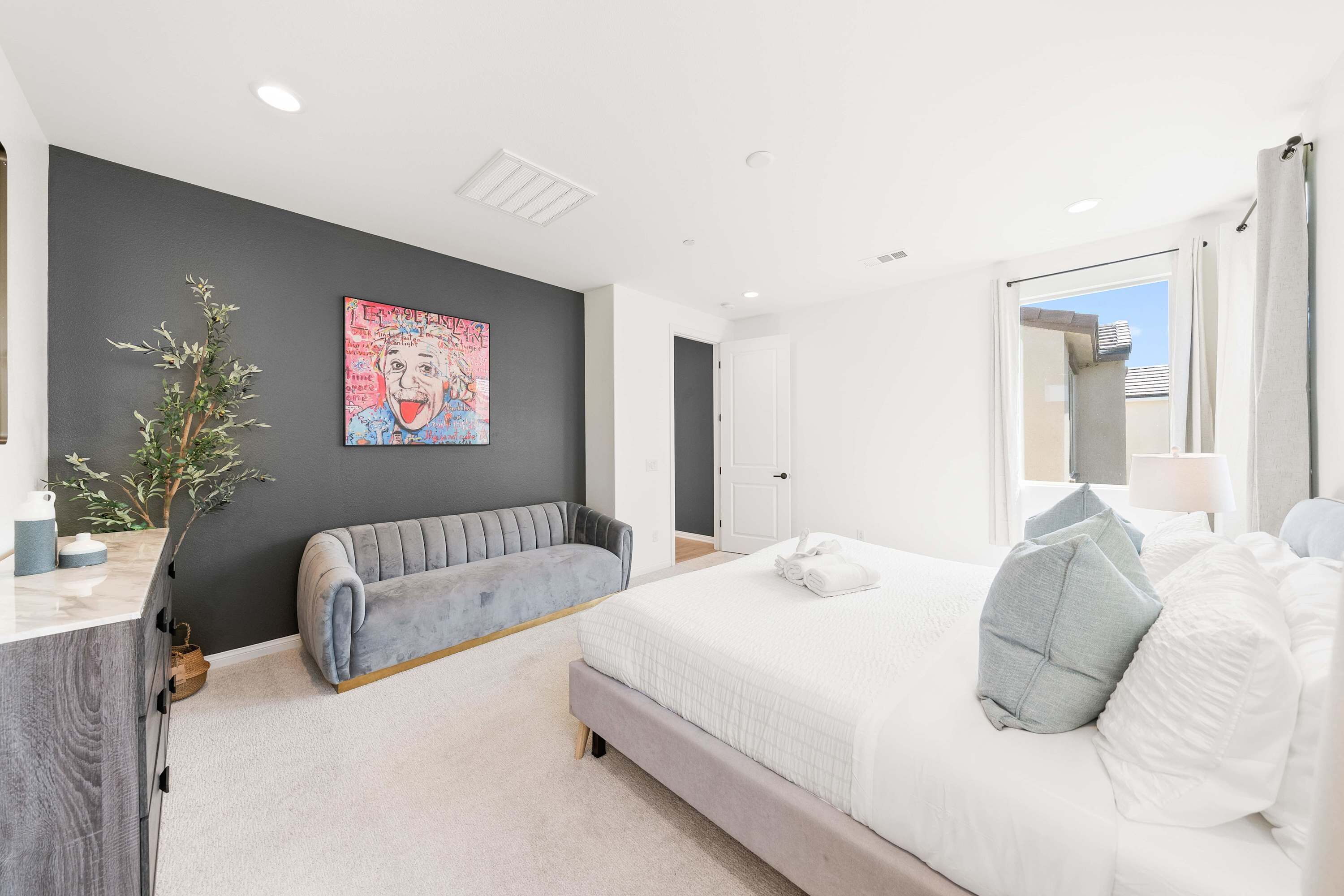 A modern bedroom with a large bed, a grey sofa, a dark accent wall, and a window with curtains.