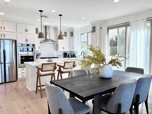 Una cocina moderna con armarios blancos, electrodomésticos de acero inoxidable y una isla central. Hay una mesa de comedor con seis sillas y un jarrón con flores.