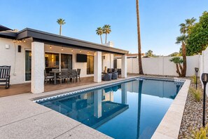 A residential swimming pool with a patio area, outdoor seating, and palm trees.