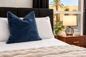 Un lit avec un matelas blanc, un oreiller bleu et un couvre-lit blanc. À côté du lit, on trouve une table de chevet en bois, sur laquelle se trouvent une lampe et une plante en pot.