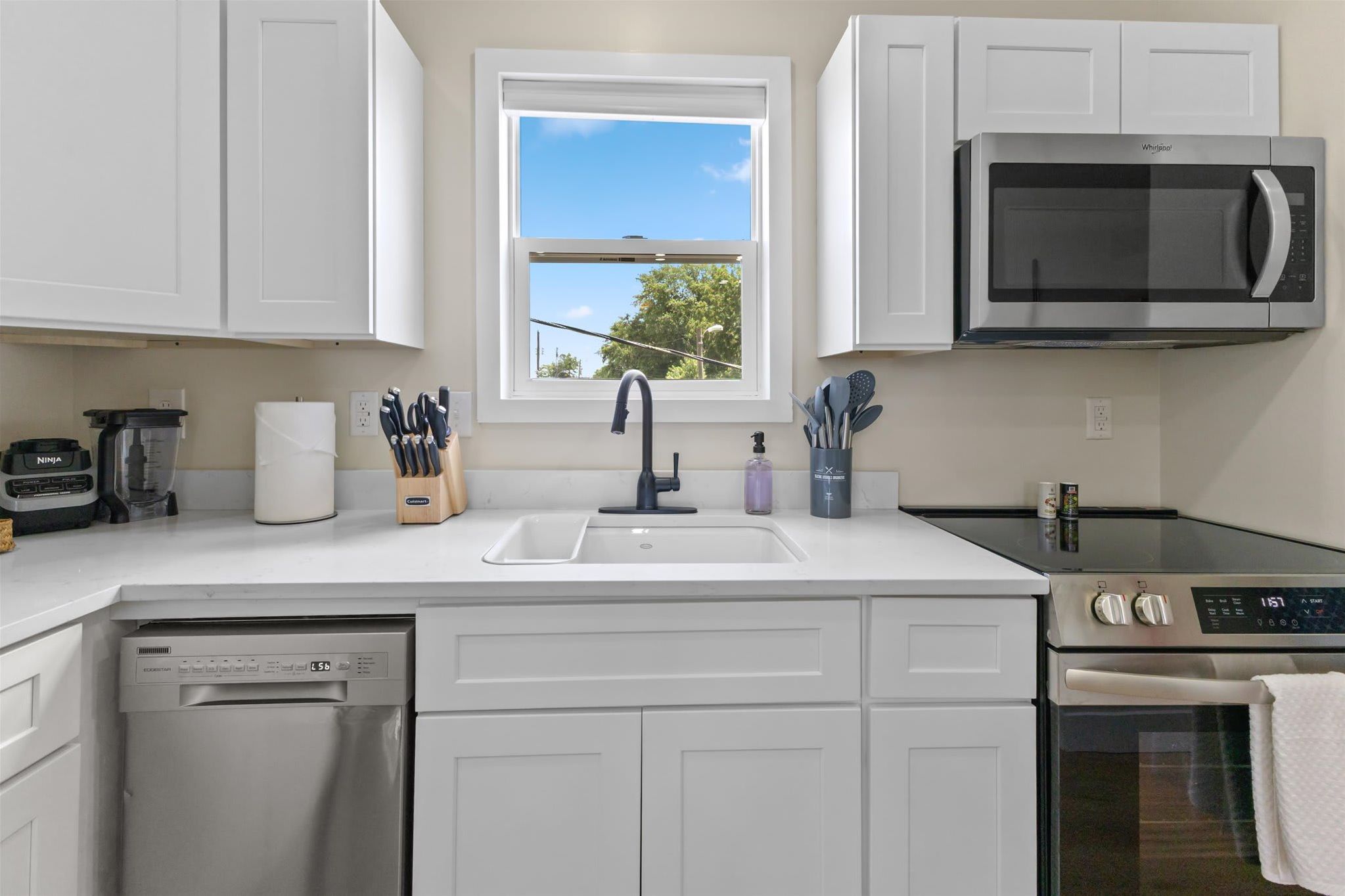 A modern kitchen with white cabinets, a built-in microwave, a dishwasher, and a window with a view of trees.