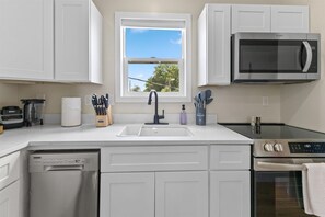 A modern kitchen with white cabinets, a built-in microwave, a dishwasher, and a window with a view of trees. - Modern Carriage House Indy (Indianapolis)