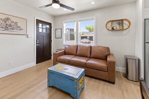 A living room with a brown leather sofa, a blue chest of drawers, a ceiling fan, and a window with a view of houses. - Modern Carriage House Indy (Indianapolis)