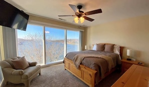 A bedroom with a large bed, a ceiling fan, a TV, and a view of a lake through large windows.