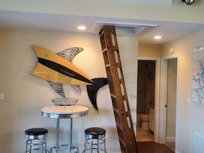A room with a round table, two stools, a decorative fish wall art, and a wooden ladder leaning against the wall.