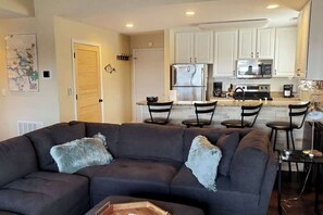 A modern living room with a dark-colored sofa and a dining area with chairs and a small table. A kitchen with white cabinets and appliances is visible in the background.