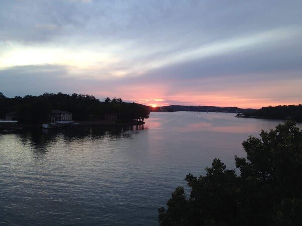 A calm lake at sunset with a house on the left and a dock extending into the water.