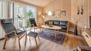 Una habitación acogedora con paredes y suelos de madera, un sofá de cuero negro, dos sillones, una mesita redonda con fruteros y una alfombra colorida.