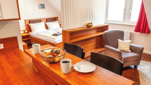 A hotel room with a wooden dining table, a bowl of pastries, two white cups, and a bed with white bedding.