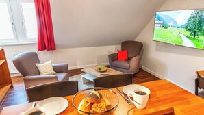 A modern living room with a wooden coffee table, two grey armchairs, a television mounted on the wall, and a window with red curtains.