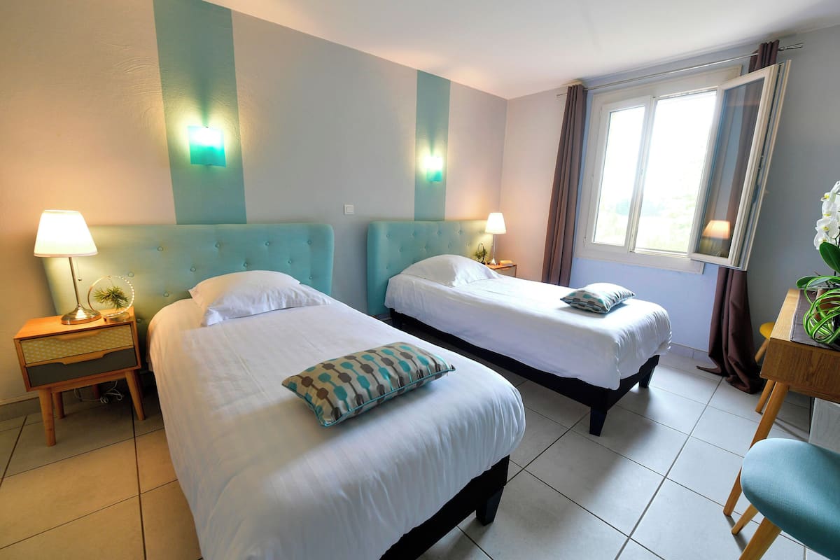 Habitación de hotel con dos camas, un escritorio y una silla. Hay mesitas de noche con lámparas y una ventana con cortinas.