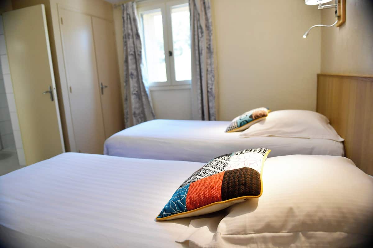 Dos camas con sábanas blancas y almohadas decorativas, una ventana con cortinas y un cabecero de madera.