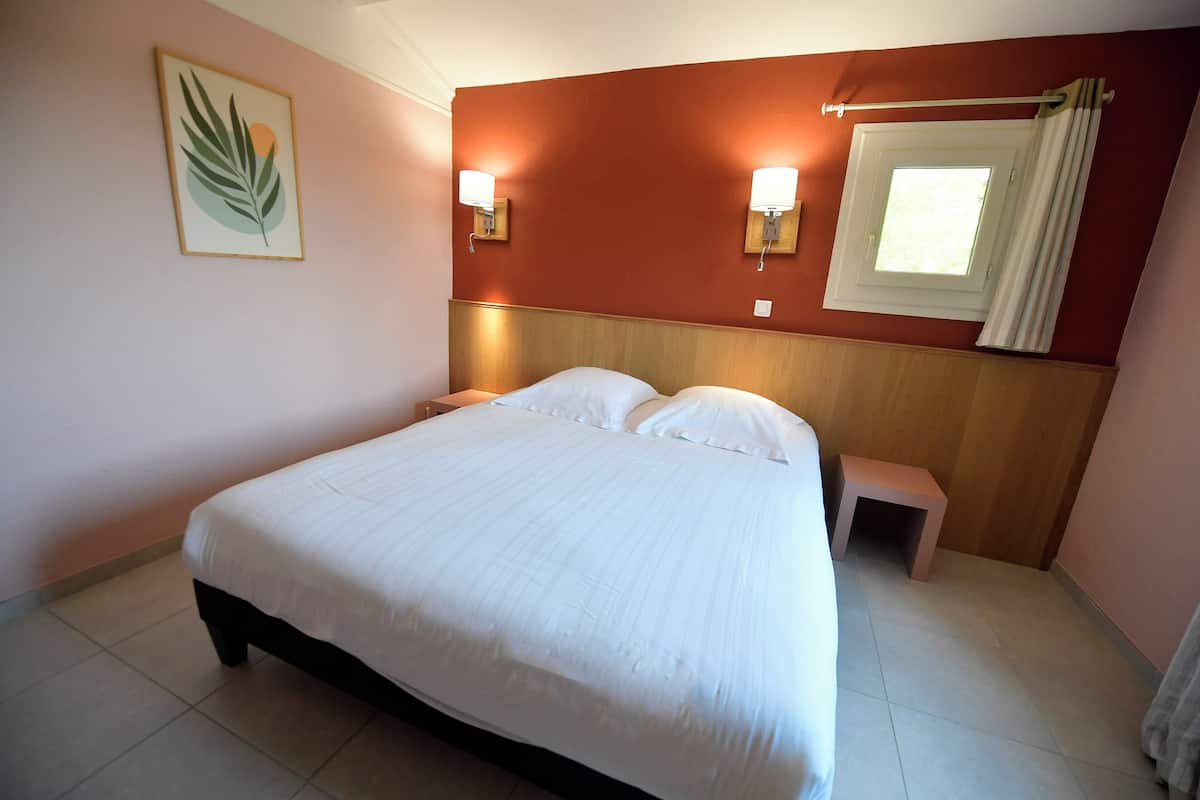 Una cama bien tendida con sábanas blancas en una habitación con cabecera de madera, dos lámparas de pared, una mesita de noche pequeña y una ventana con cortinas.
