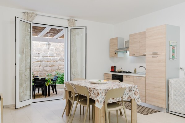 A modern kitchen with wooden cabinets, a dining table with chairs, and a view through an open door to a stone wall and plants. - Apartment 'Mea' with Sea View, Wi-Fi and Air Conditioning (Balestrate)