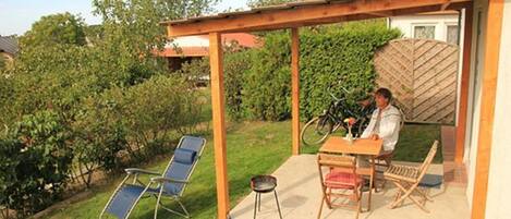 A covered patio with a table and chairs, surrounded by greenery and a wooden fence.