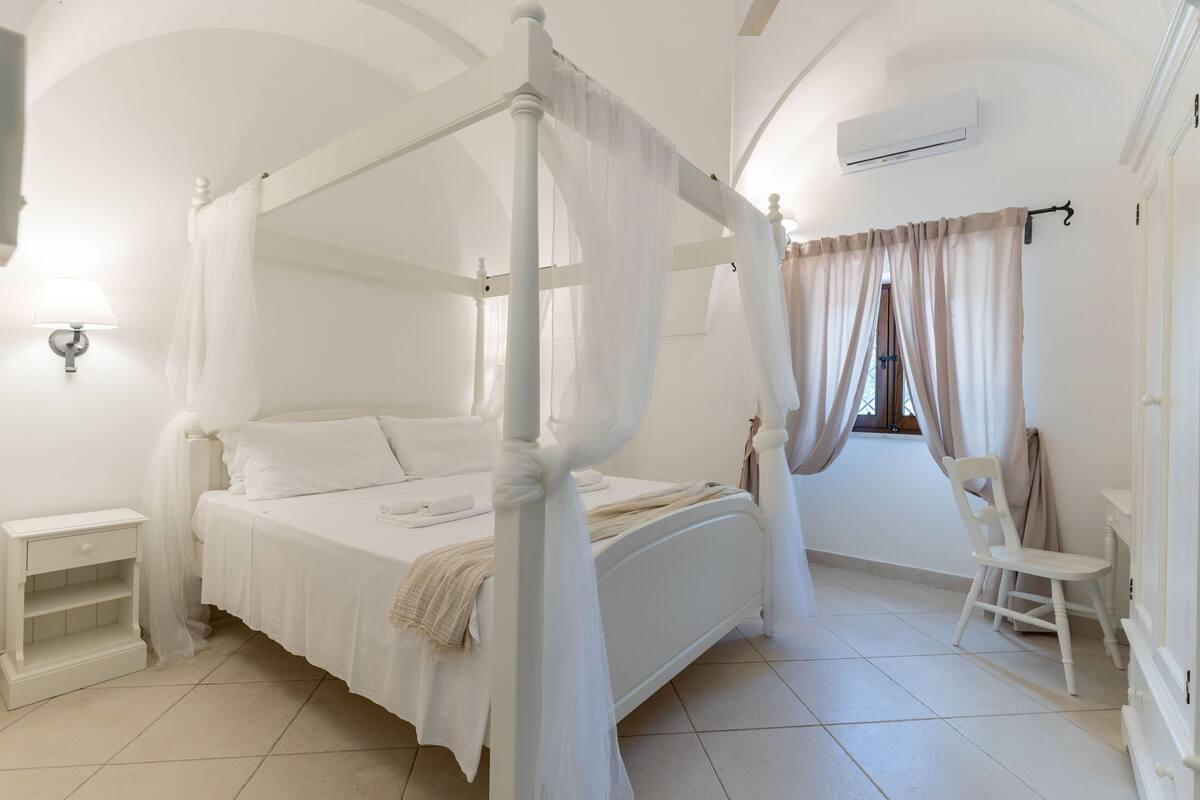 Ein Himmelbett mit weißem Finish, ein Nachttisch, ein Sessel und ein Fenster mit Vorhängen.