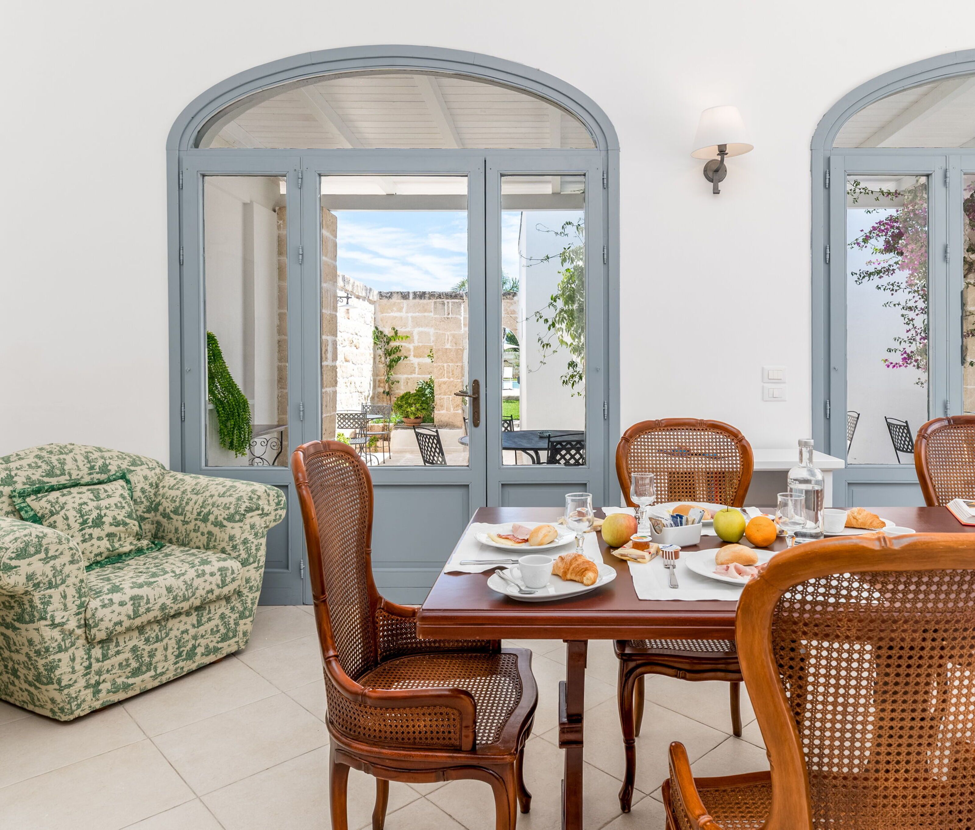A dining area with a table set for two, surrounded by wicker chairs, with a view of the courtyard through arched windows.