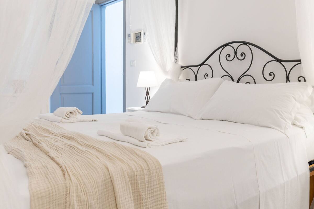 Ein ordentlich bezogenes Bett mit weißen Leinentüchern und einer beige farbenen Tagesdecke, ein schwarzes Metall-Kopfteil und eine blaue Tür im Hintergrund.