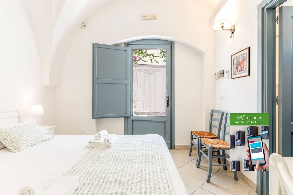 Ein Hotelzimmer mit Bett, Sessel, Schreibtisch, Lampe, einem Fenster mit Vorhängen und einer Tür mit Schild für Selbstcheck-in und intelligenten Zugang.