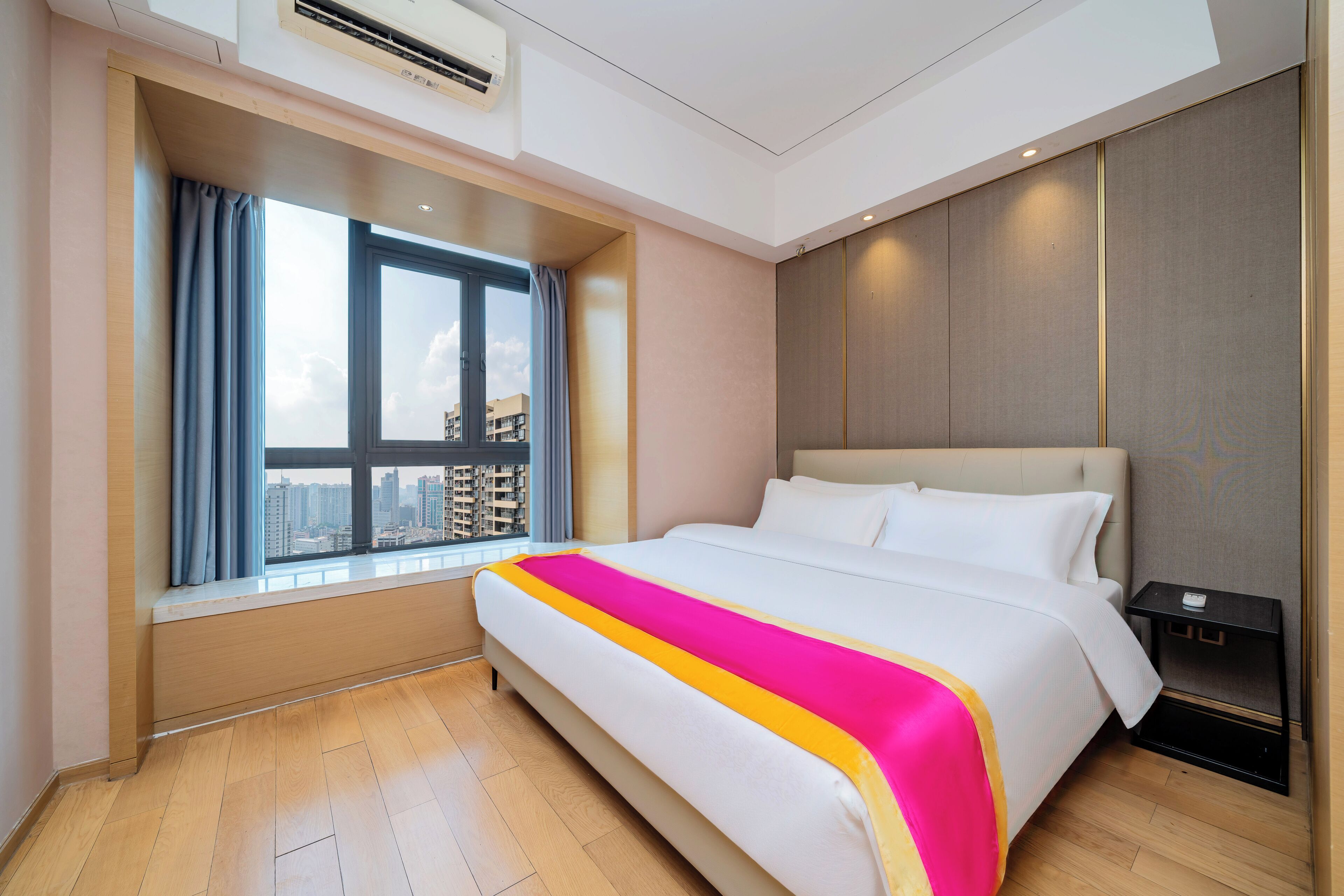 Une chambre d’hôtel avec un grand lit, une fenêtre donnant sur des immeubles, une table de chevet et un climatiseur fixé au plafond.