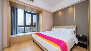 A hotel room with a large bed, a window with a view of buildings, a nightstand, and a ceiling-mounted air conditioner.