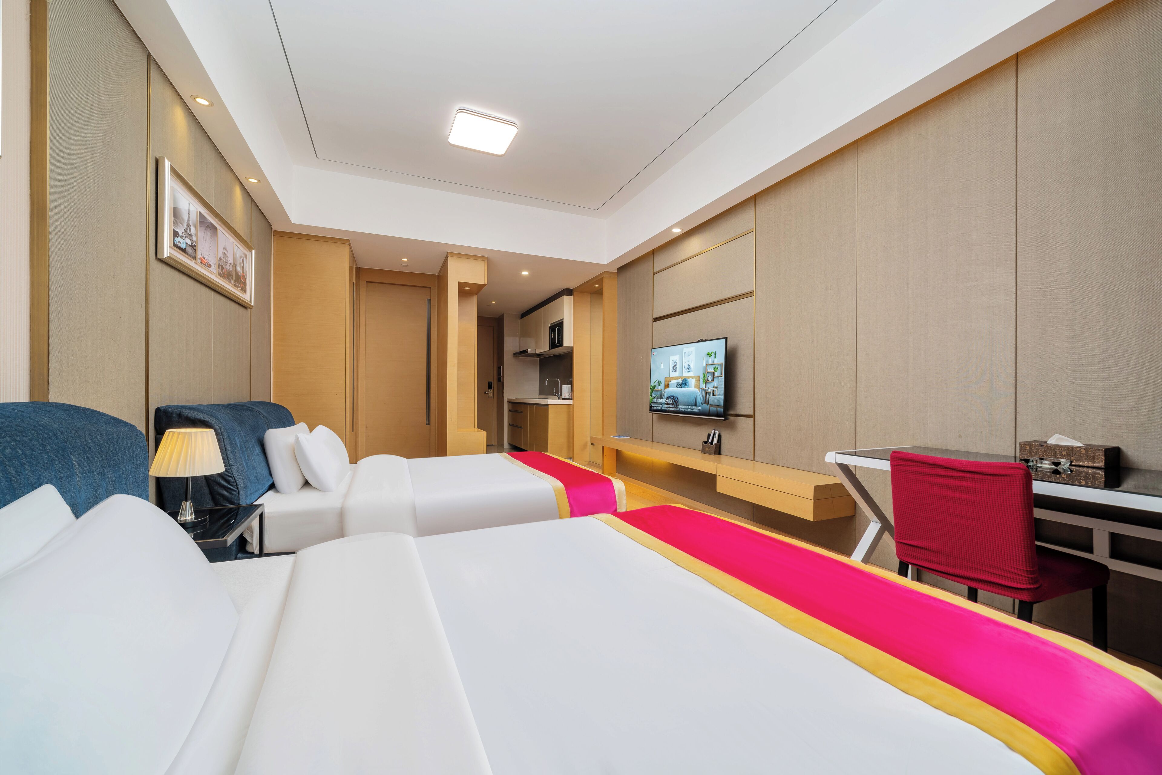 Habitación de hotel con dos camas, un escritorio, un televisor y una kitchenette pequeña.