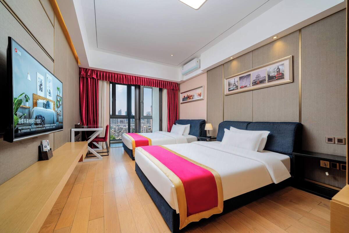 A hotel room with two beds, a TV, a desk, and a view of the city.