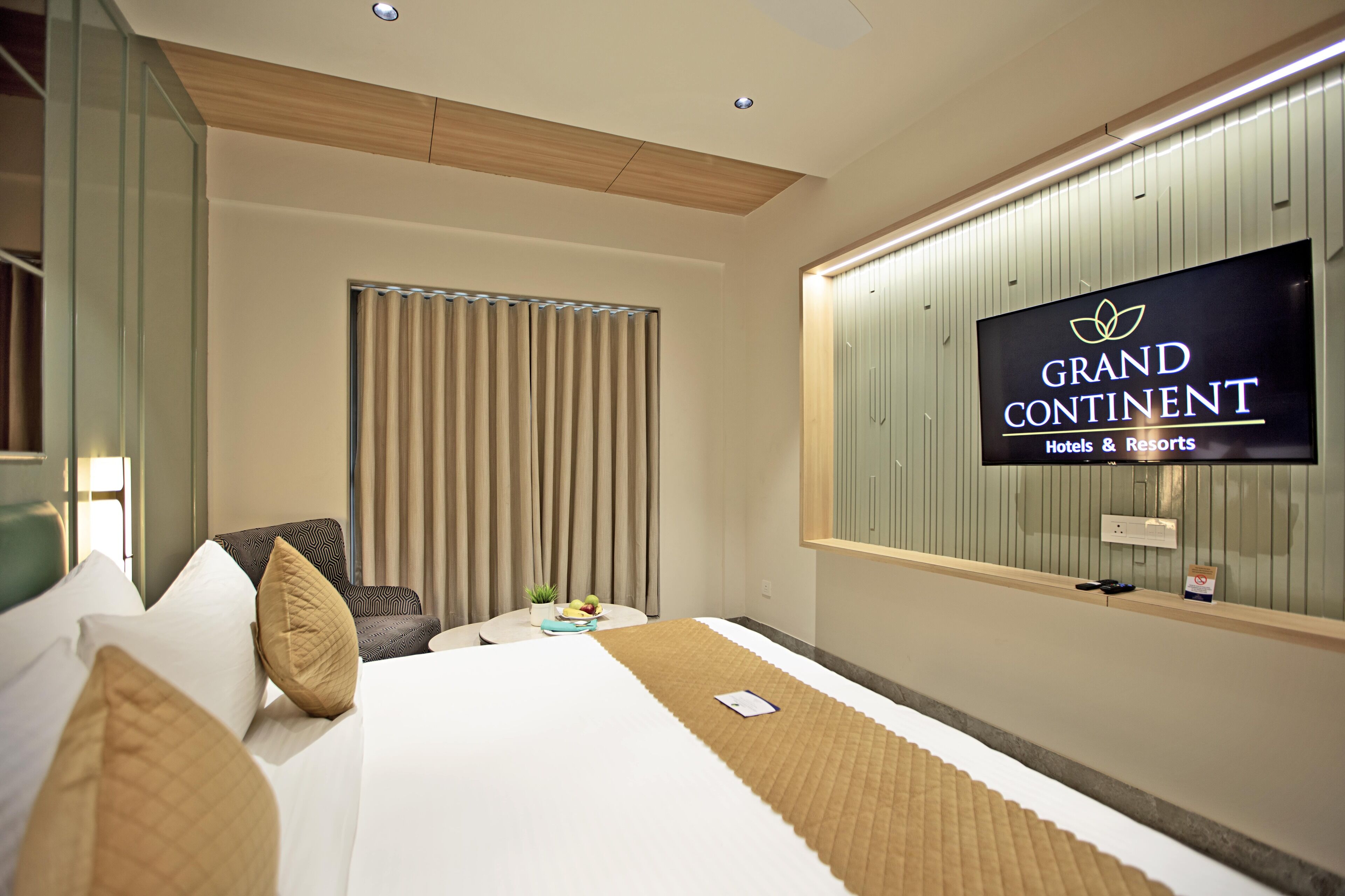 Una habitación de hotel con cama, televisión montada en la pared, ventana con cortinas y una pequeña mesa con una planta y fruta.