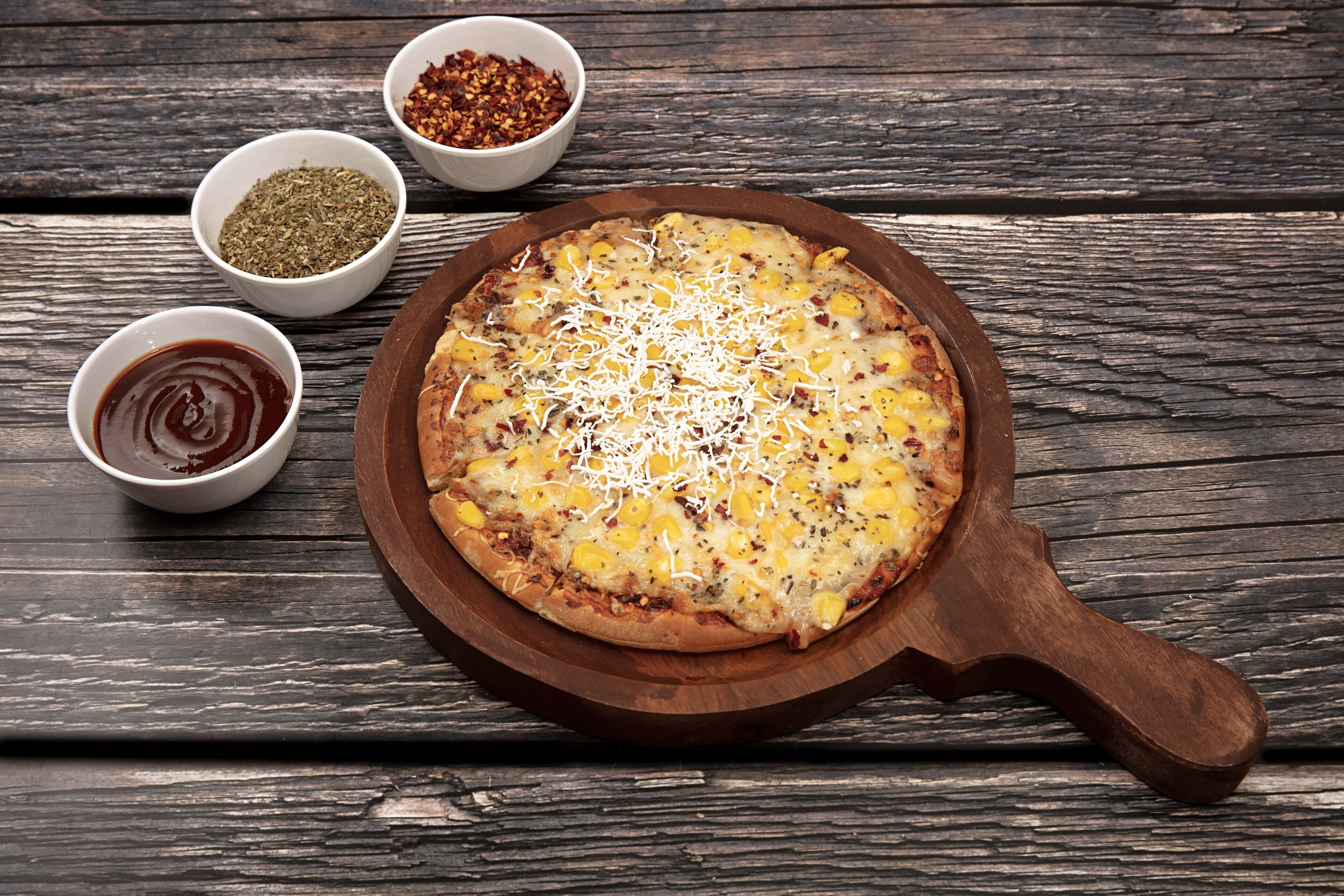 Una pizza sobre una tabla de madera con asa, acompañada de recipientes pequeños con salsas y condimentos.
