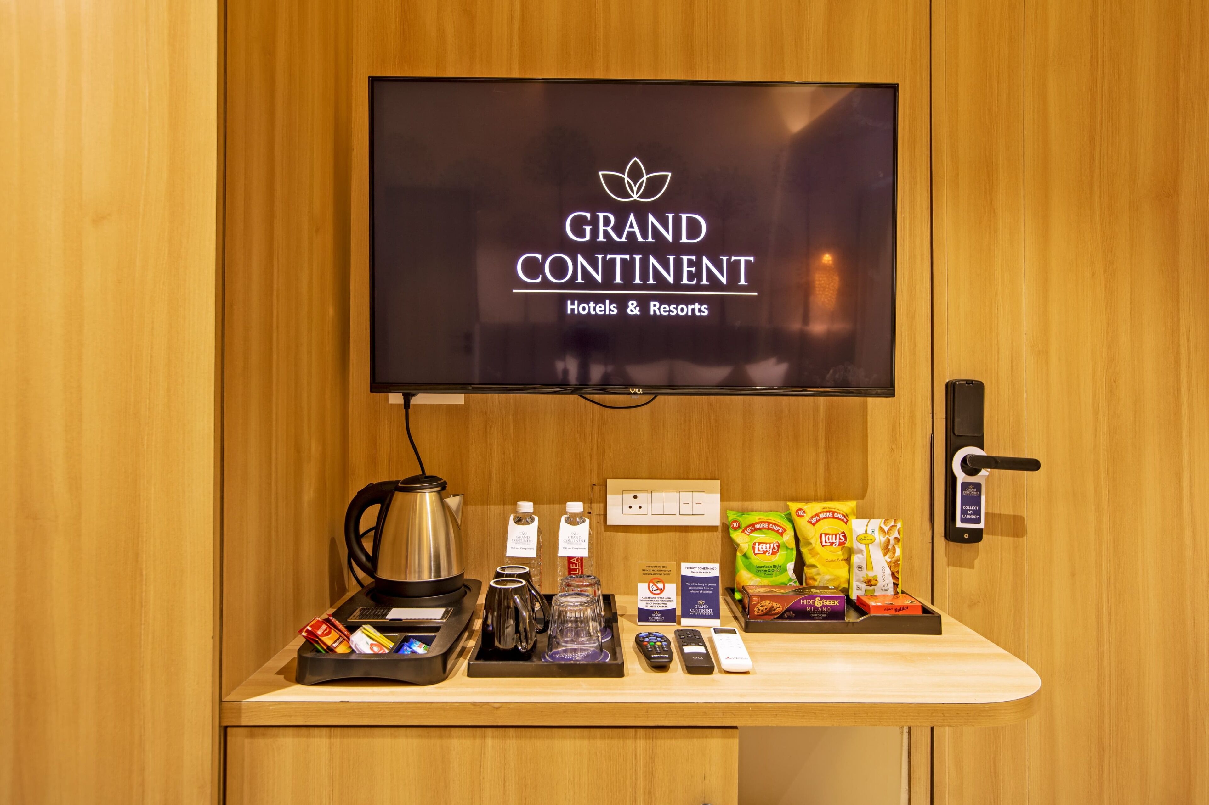 A hotel room with a television displaying 'GRAND CONTINENT Hotels & Resorts' logo, a coffee maker, a kettle, a tray with snacks, and a remote control.