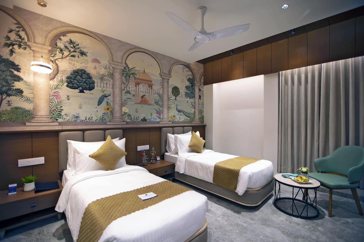 A hotel room with two beds, a small table, a chair, a ceiling fan, and a mural on the wall.