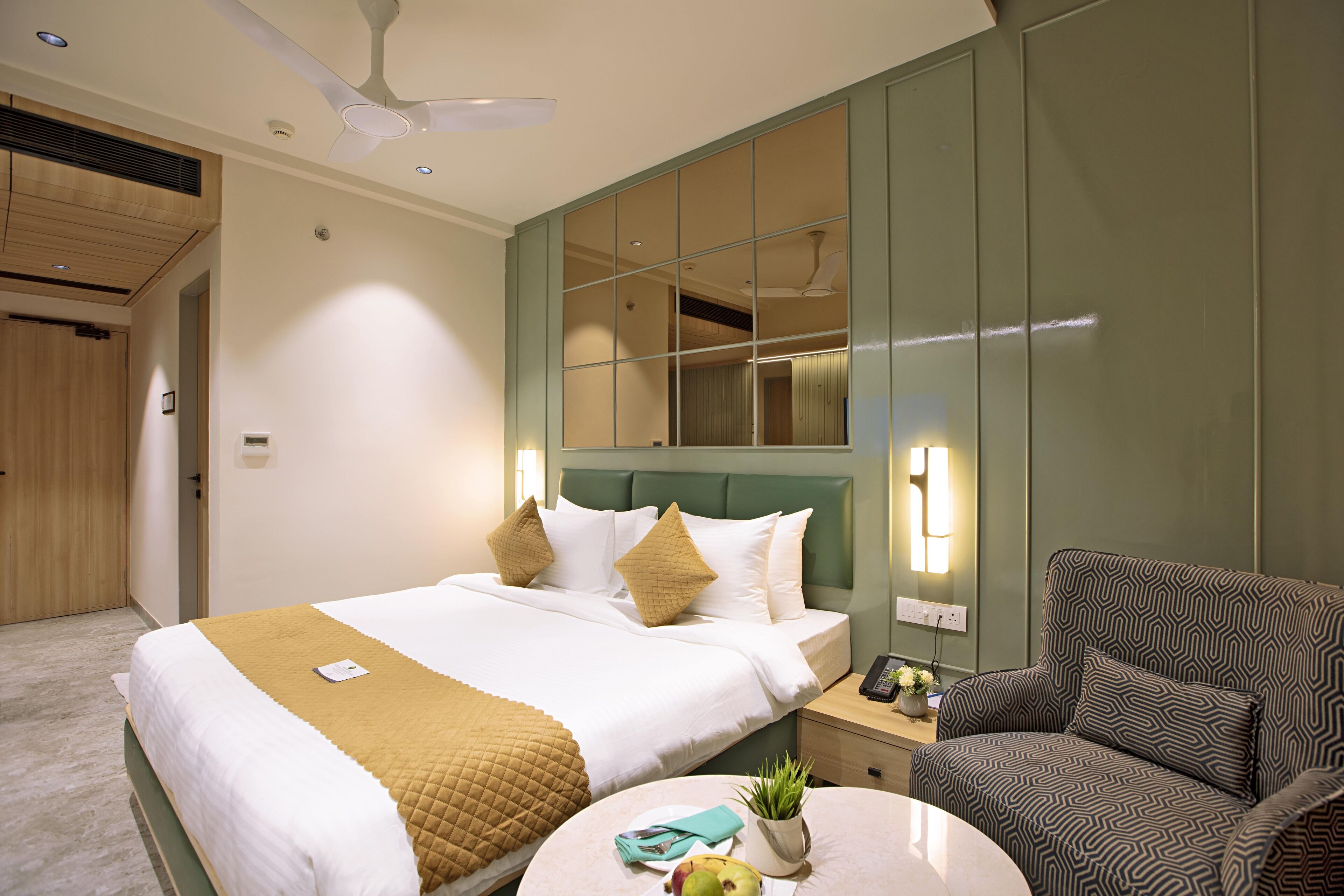 A hotel room with a bed, bedside table, chair, and a round table with a plant and fruit.