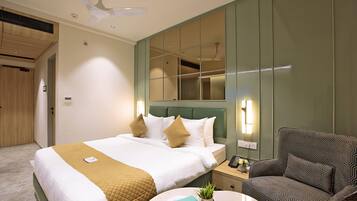A hotel room with a bed, bedside table, chair, and a round table with a plant and fruit.